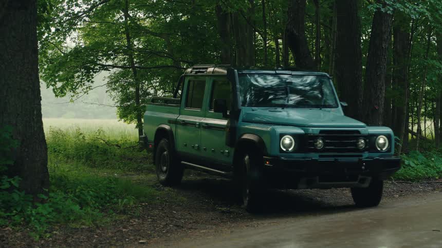 A teal off-road pickup truck emerges from the woods and drives down a wet dirt road on an overcast day