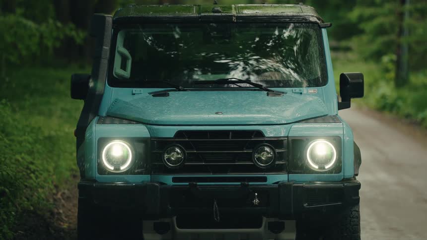 Frontal view of a modern teal off-road vehicle with headlights on and windshield wipers operating during a downpour in a lush green forest