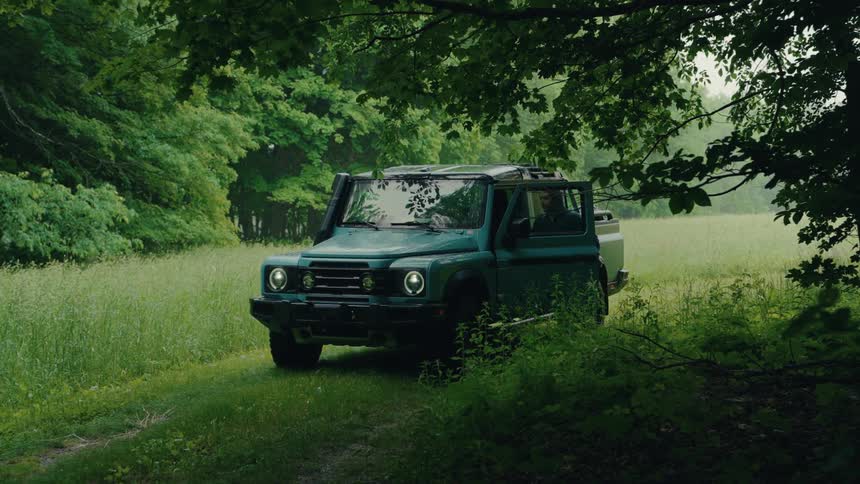 A man gets into the driver's seat of a green 4x4 truck parked on a grassy trail in a lush forest