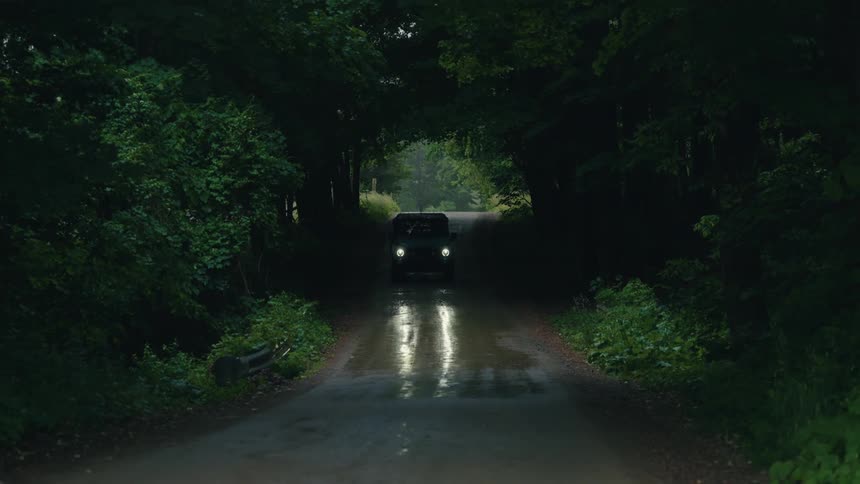 An off-road vehicle with headlights on slowly emerges from a dark tree tunnel on a wet country road