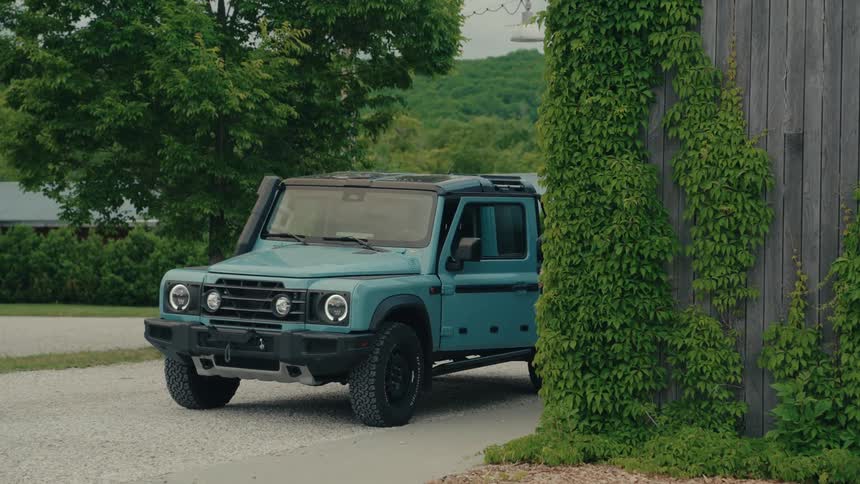 A young man gets out of a modern blue SUV and walks away from it in the driveway of an ivy-covered country home