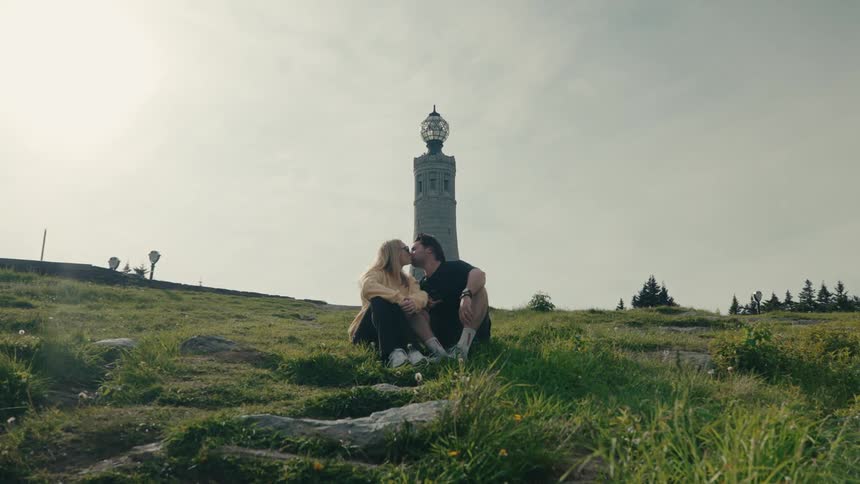 A man walks up a grassy hill in slow motion to meet his partner, and they share a romantic kiss and embrace in front of a tall stone monument