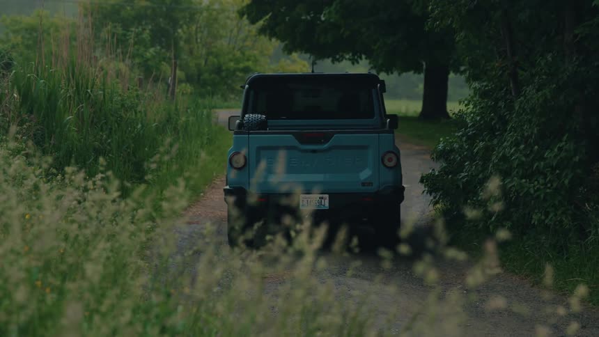 A modern blue off-road pickup truck reverses slowly down a winding dirt road in the country