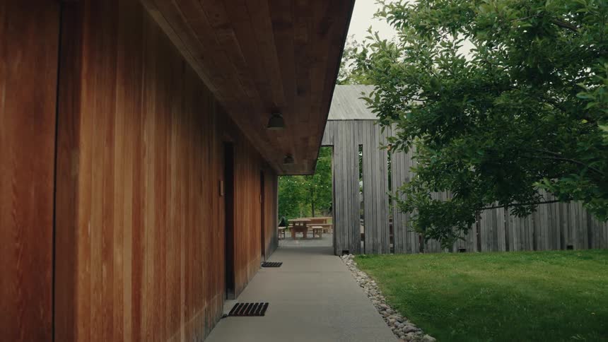 A quiet concrete pathway runs alongside a modern wooden building next to a green lawn and trees