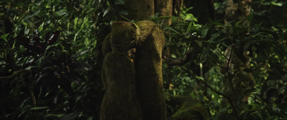 A moss-covered stone sculpture of two intertwined lizards stands amongst the dense foliage of a lush jungle