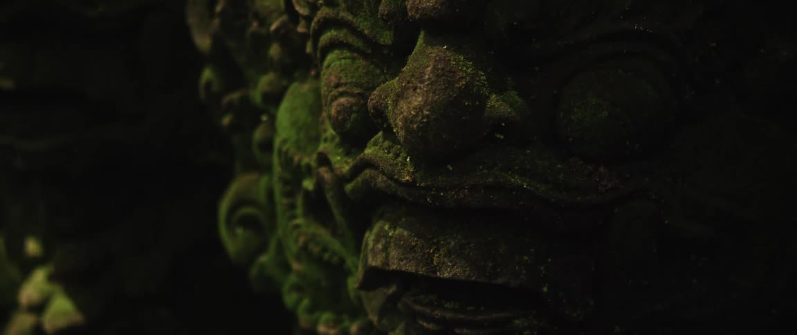 A close-up slow focus pull reveals the weathered, moss-covered face of an ancient stone temple carving