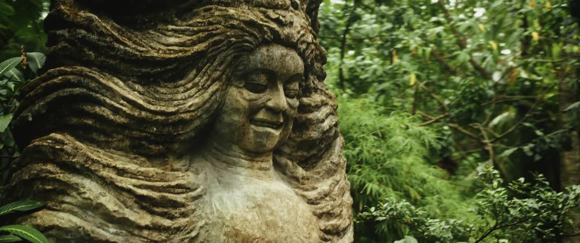 A mysterious and weathered stone statue of a smiling woman with flowing hair is slowly revealed in a lush jungle garden