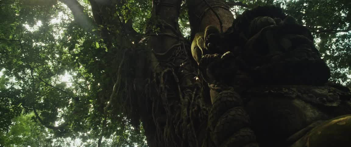 An ancient, moss-covered statue of a deity stands guard under a massive banyan tree in a mystical jungle