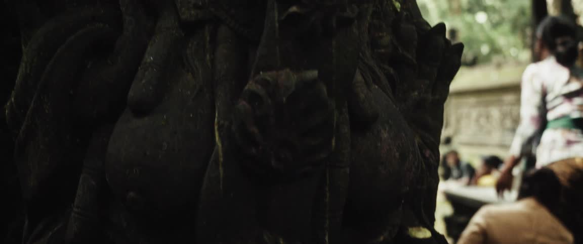 Slow tilt-up reveals a moss-covered ancient stone statue of a Balinese deity, starting with a carved skull