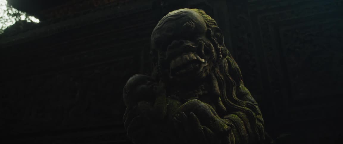 An ominous low-angle shot of a moss-covered ancient stone statue depicting a demon holding a child