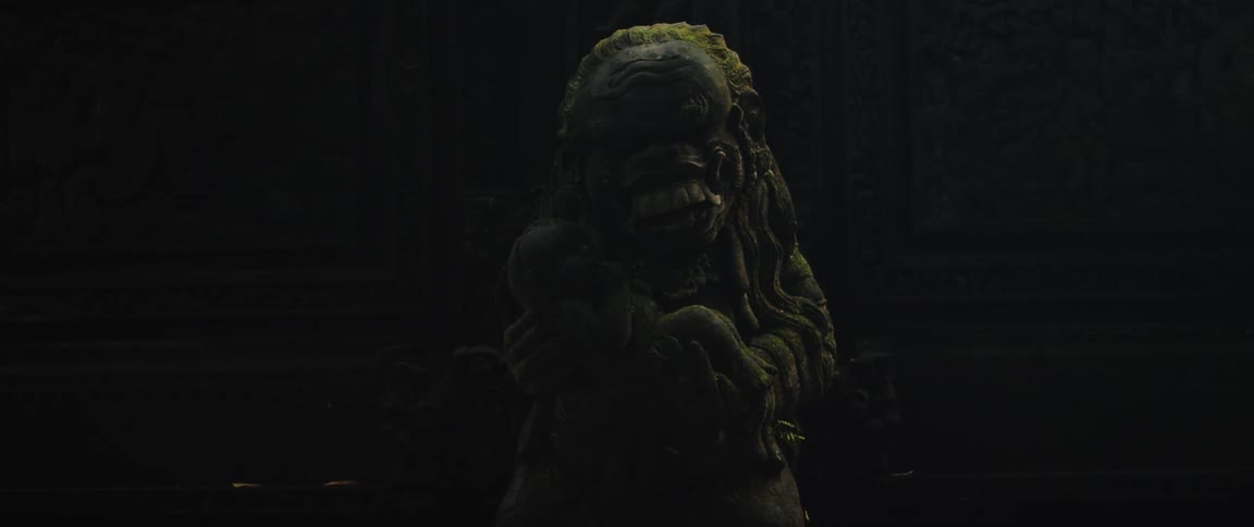 An eerie, moss-covered stone statue of a mythological creature holding a baby is dimly lit in an ancient temple