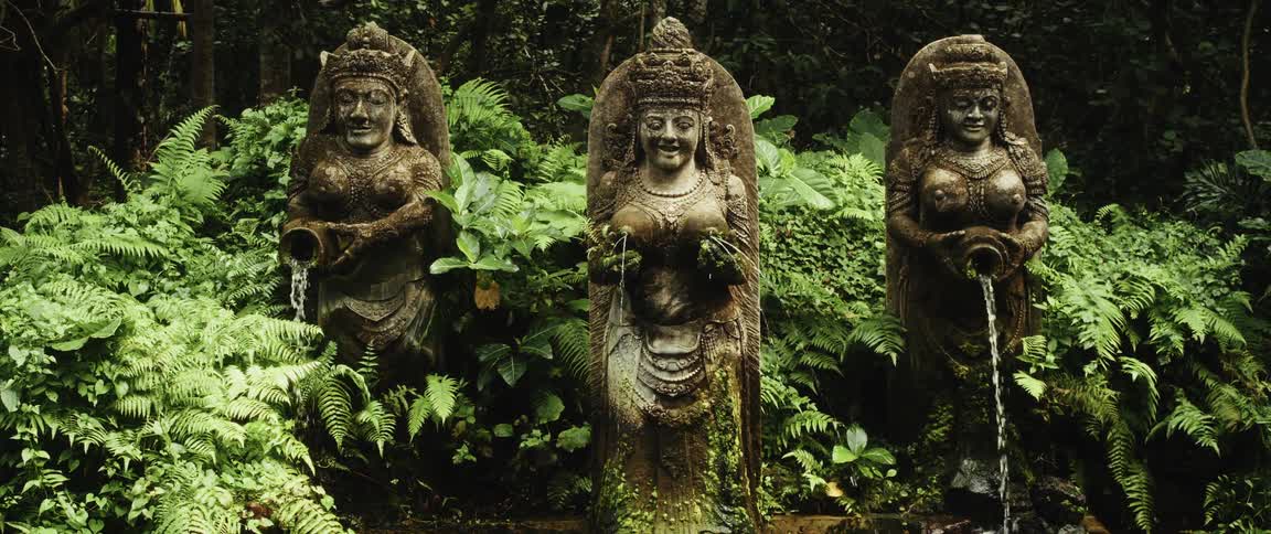 Water flows from three serene, ancient statues of female deities surrounded by dense green foliage