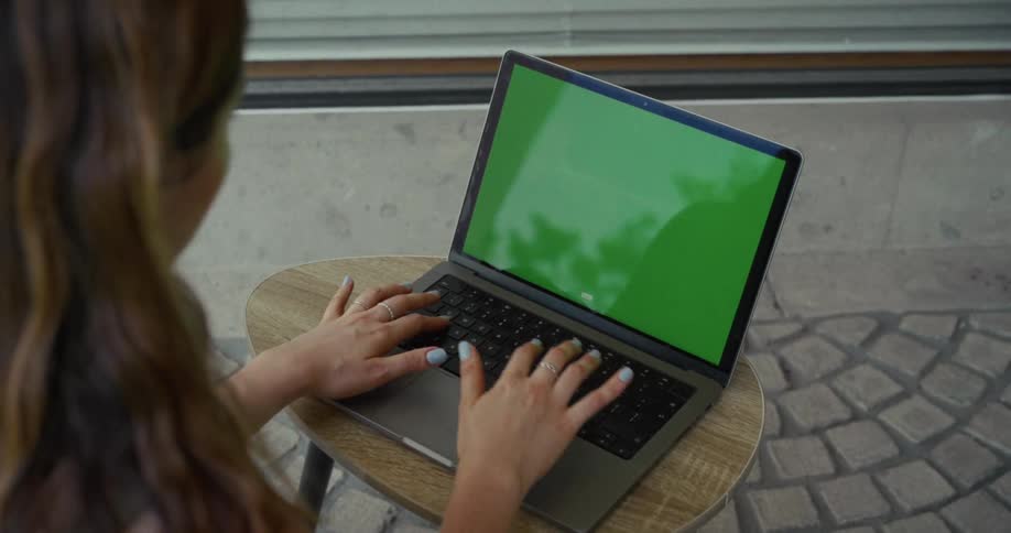 An over-the-shoulder shot of a woman typing on a laptop with a green screen outdoors on a stone patio