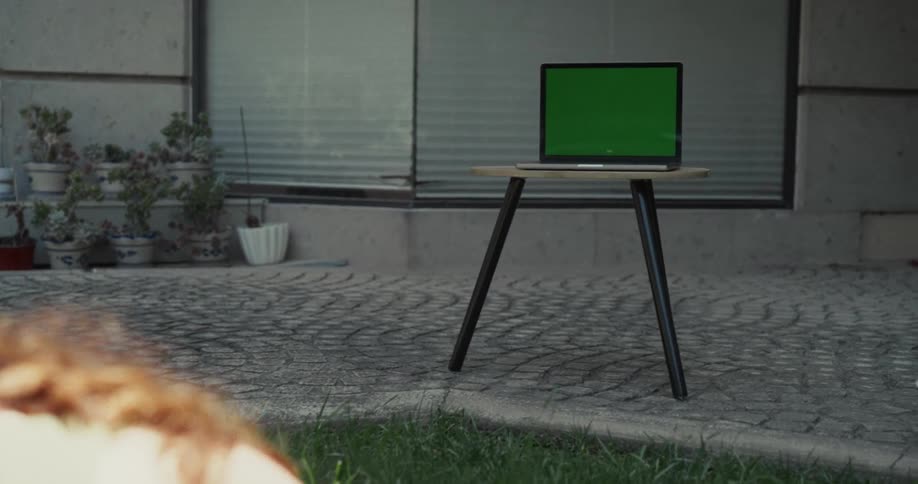 A slow push-in on a green screen laptop as a woman in the foreground follows an online yoga class in her backyard
