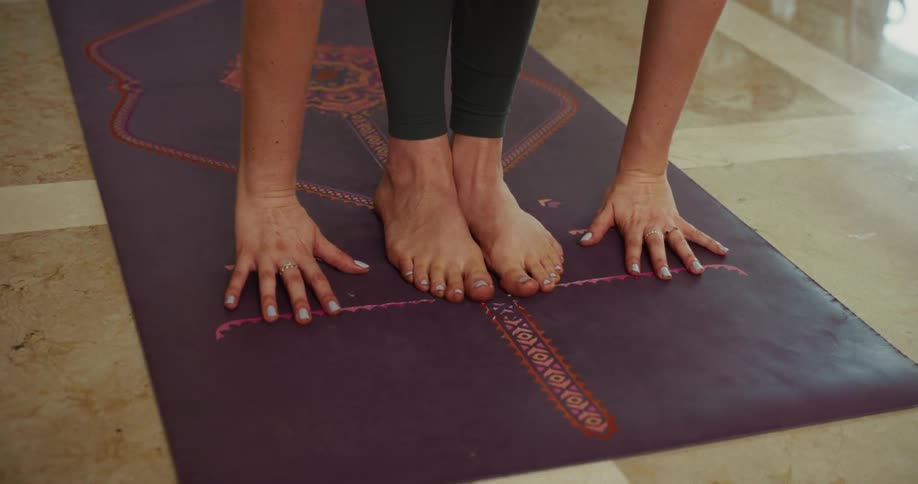 A woman's hands and feet rest on a purple yoga mat as she performs a standing forward bend