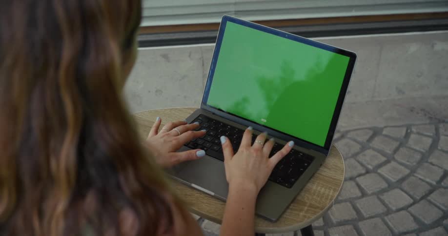 Over-the-shoulder view of a woman with long brown hair typing on a laptop with a green screen at a small table on a stone patio
