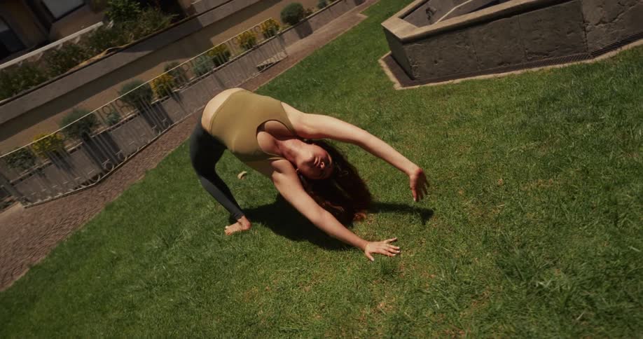 High-angle shot of a woman gracefully performing a deep yoga backbend on a green lawn in slow motion