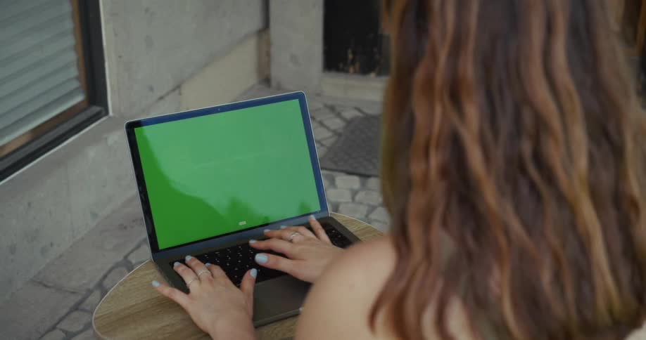Over the shoulder shot of a young woman typing on a modern laptop with a green screen while sitting at an outdoor cafe table