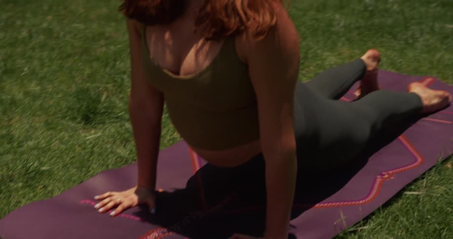 A serene young woman with red hair practices a yoga flow on a purple mat in a sunny backyard, transitioning from downward dog to a peaceful upward-facing dog