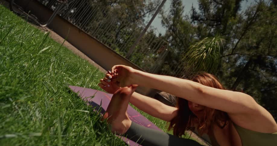 A flexible woman performs a deep forward bend stretch while practicing yoga on a mat in her lush, sunny backyard