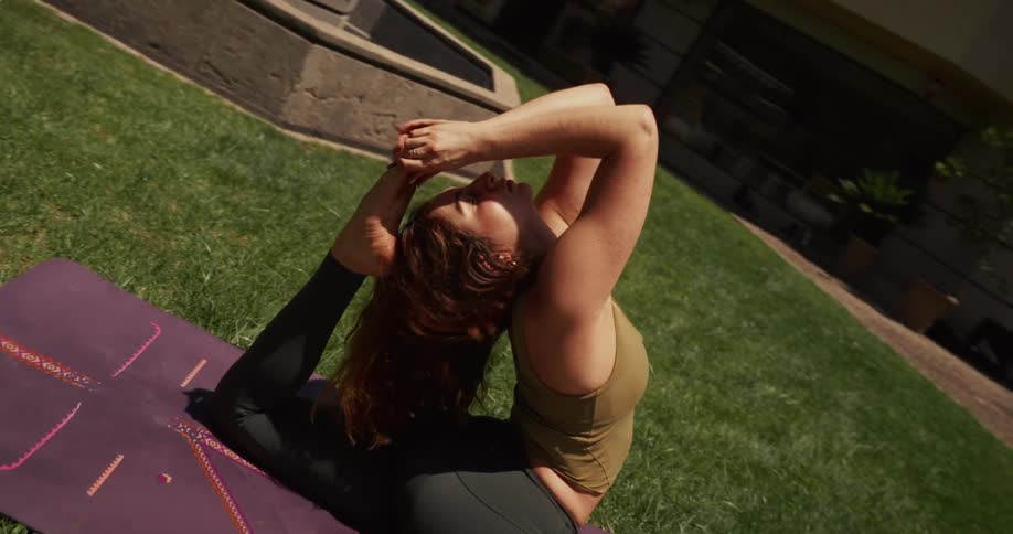 A flexible woman in a dutch angle shot holds an advanced yoga pose on a purple mat in a sunny backyard