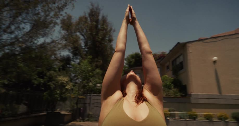 Slow motion shot of a woman in a tranquil backyard, gracefully raising her arms into a prayer pose while practicing yoga under the sun