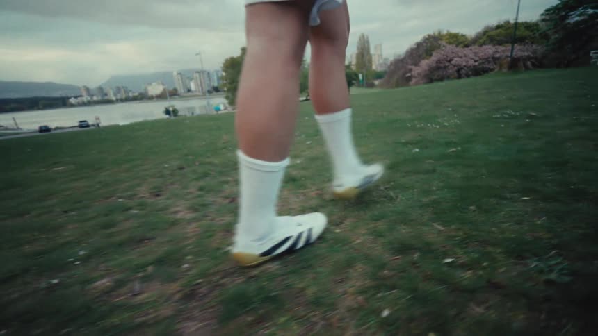 A slow-motion, low-angle shot follows a soccer player's cleats as they walk across a grassy park with a city waterfront in the background