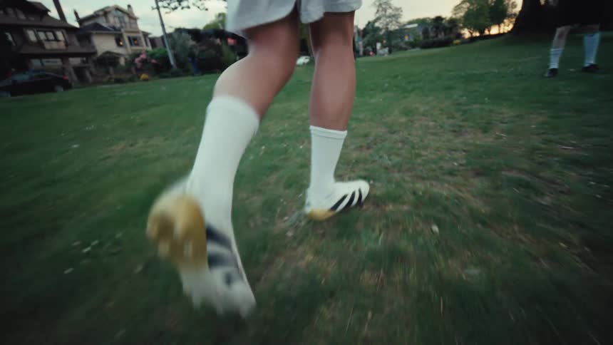 A close-up slow-motion shot follows a soccer player's feet as they dribble a ball across a grassy park field at dusk