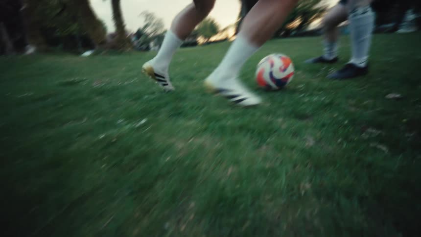 A dramatic low-angle slow-motion shot follows a soccer player's feet as they skillfully dribble a ball across a grassy park field