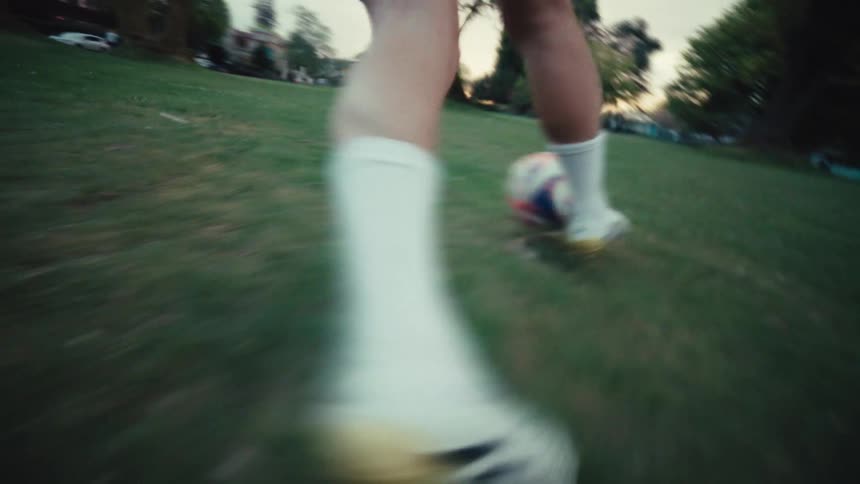 A slow-motion, low-angle shot of a soccer player's feet in cleats as they dribble a ball across a grassy park field