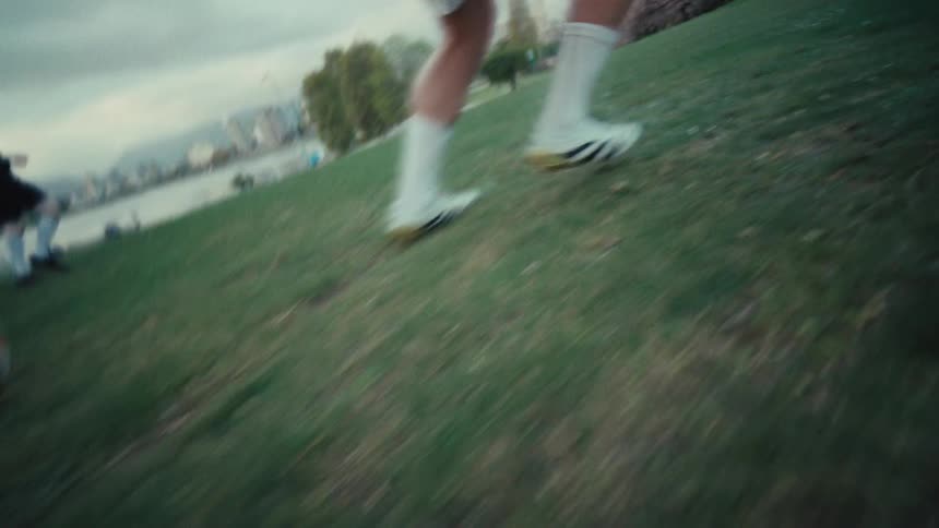 A dynamic, low-angle slow-motion shot follows a soccer player's feet as they dribble a ball across a grassy park
