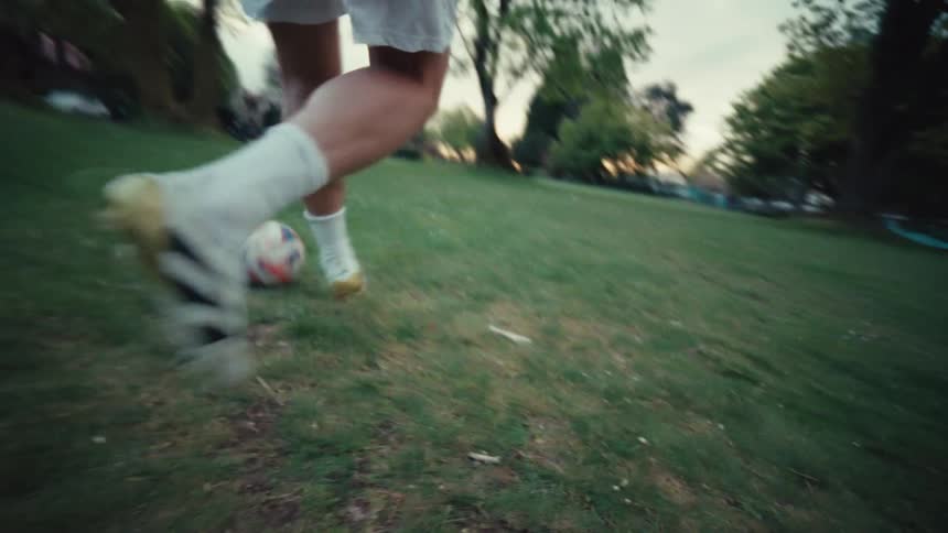 A low-angle slow-motion shot follows a soccer player's feet and cleats as they skillfully dribble the ball across a grassy park field