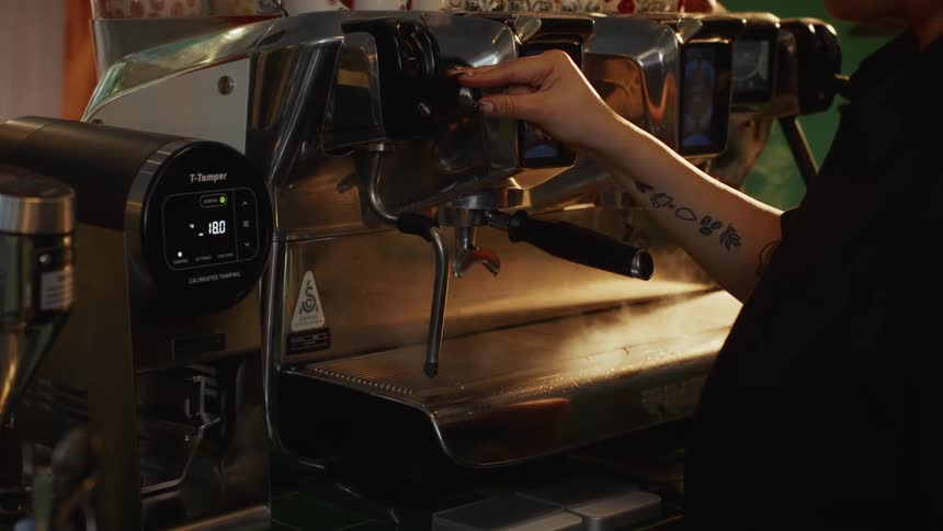 A tattooed barista releases a cloud of hot vapor from a professional espresso machine's steam wand in a cafe