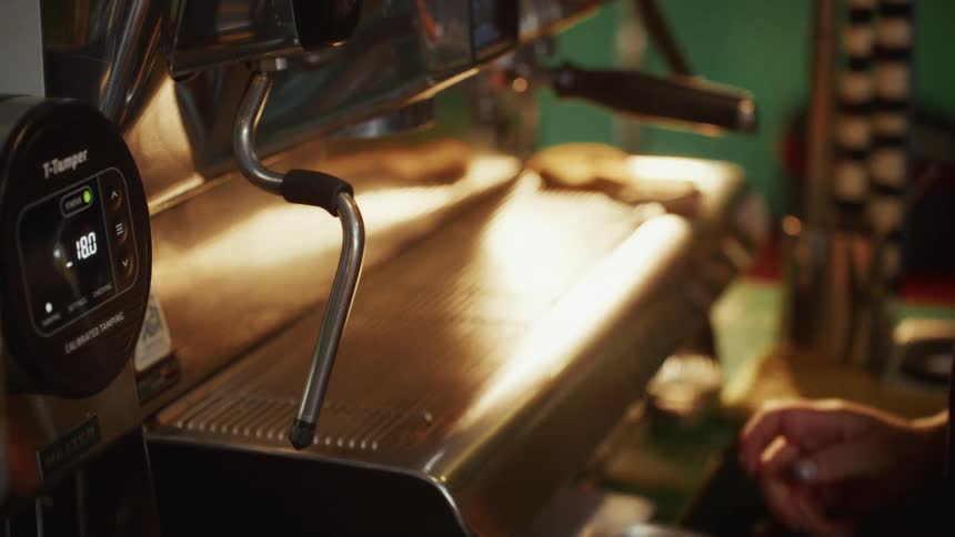 A barista prepares a portafilter while another barista cleans the espresso machine in a warmly lit coffee shop