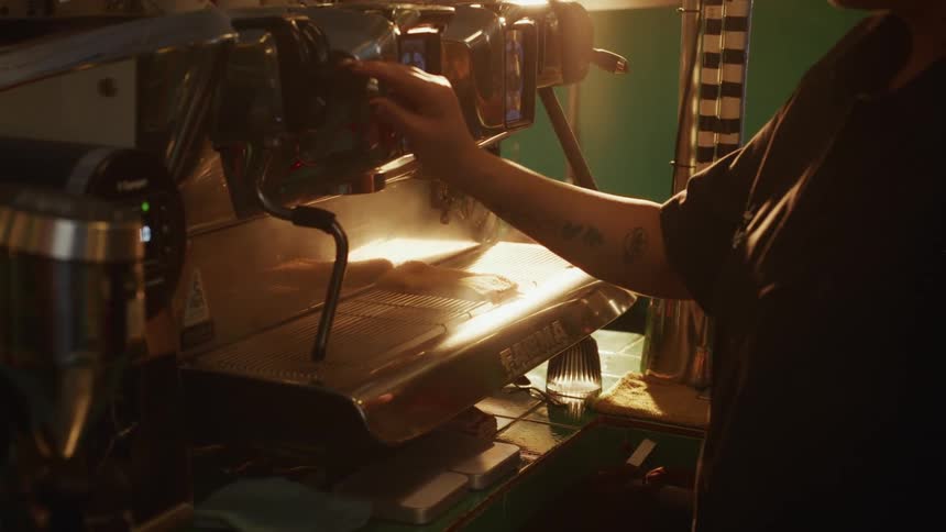 A barista with a tattoo on their arm releases steam from a chrome espresso machine in the warm light of a coffee shop