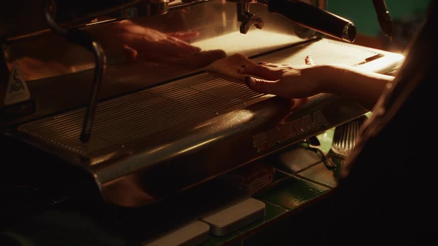 A barista carefully wipes down a stainless steel espresso machine in the warm, atmospheric light of a cafe
