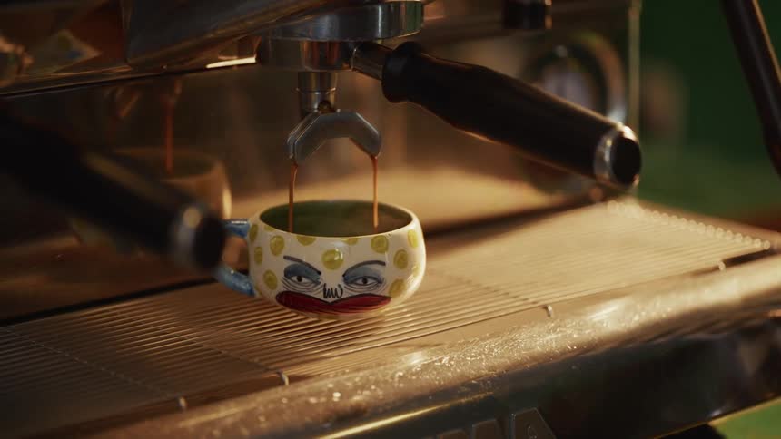 A professional espresso machine pours hot, steaming coffee into a quirky, handmade ceramic mug with a grumpy face