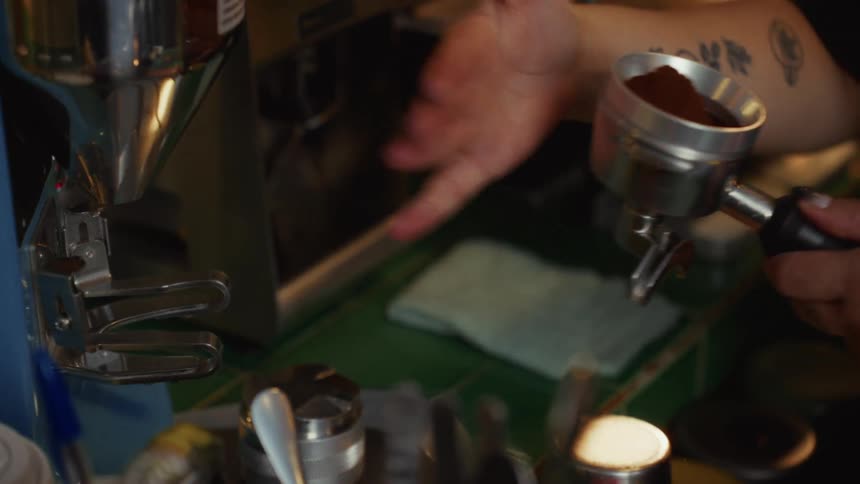 A tattooed barista grinds fresh coffee beans and carefully levels the grounds in a portafilter in slow motion