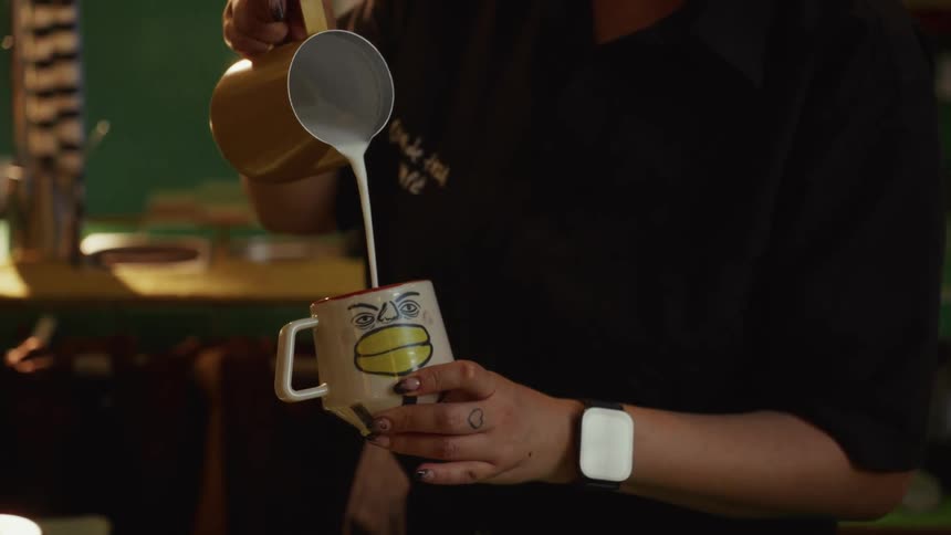 Close-up of a barista carefully pouring steamed milk into an artistic, face-painted coffee cup in slow motion