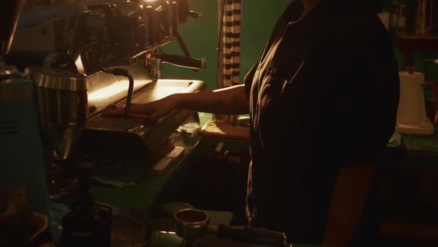 A barista purges the steam wand on a professional espresso machine in a dimly lit, cozy coffee shop