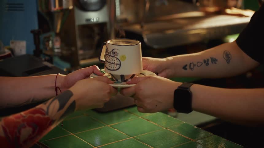 A barista hands a steaming cup of coffee in a quirky mug to a tattooed customer over a green tiled counter