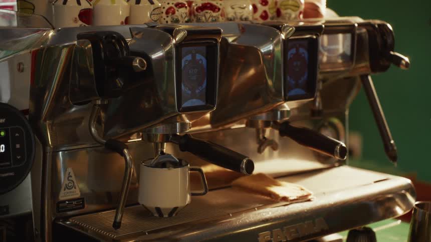 A chrome espresso machine pours a fresh shot of coffee into a white mug in a sunlit cafe
