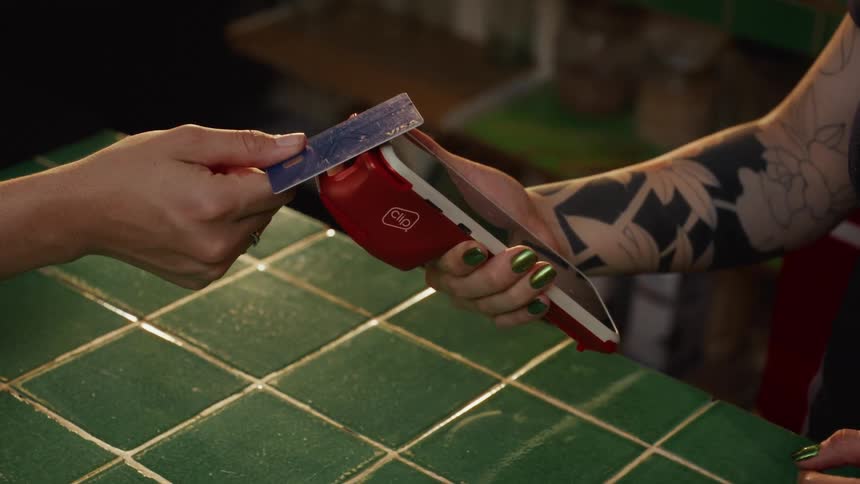 A close-up of a customer swiping a credit card through a mobile payment terminal held by a tattooed cashier