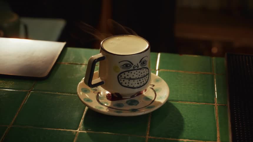A quirky mug with a hand-painted grinning face steams with a hot beverage on a green tiled counter