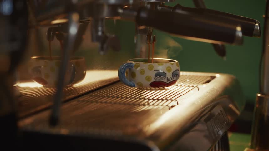 Steaming espresso pours from a coffee machine into a whimsical hand-painted ceramic mug with a tired-looking face