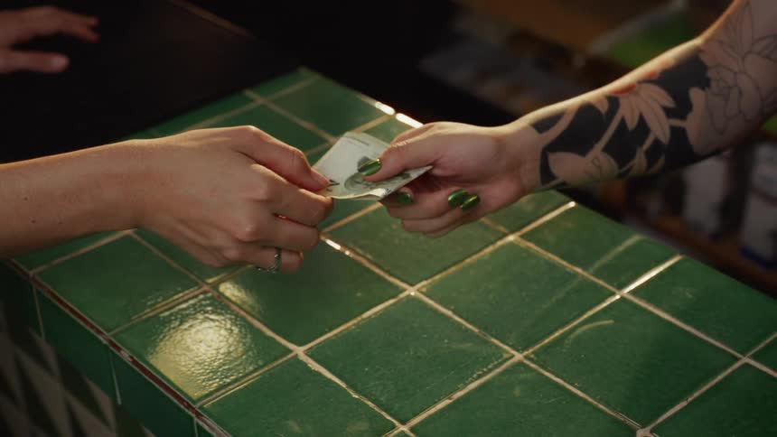 A close-up slow-motion shot captures a customer handing cash to a tattooed cashier with green nails over a green tiled counter