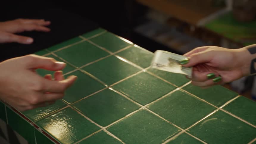 A customer pays a cashier with a cash banknote over a green tiled counter in slow motion