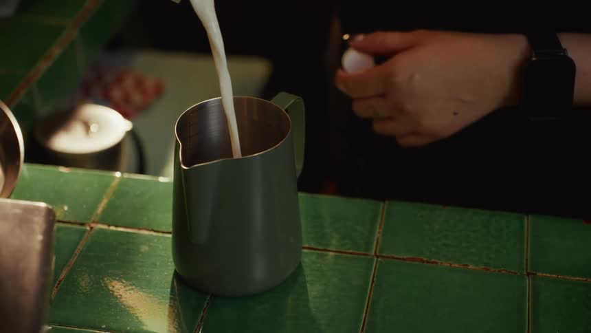 A barista pours milk into a stainless steel frothing pitcher on a green tiled counter in slow motion