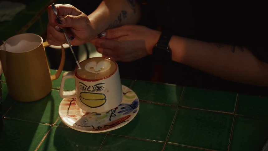 A barista carefully creates a heart-shaped latte art design in a quirky, hand-painted mug on a green tiled counter