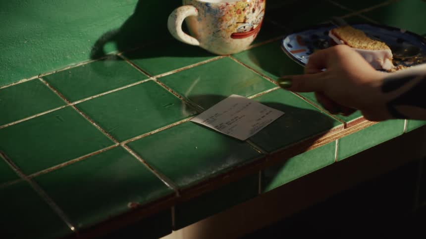 A tattooed person places a receipt down on a sunlit, green-tiled kitchen counter next to breakfast leftovers